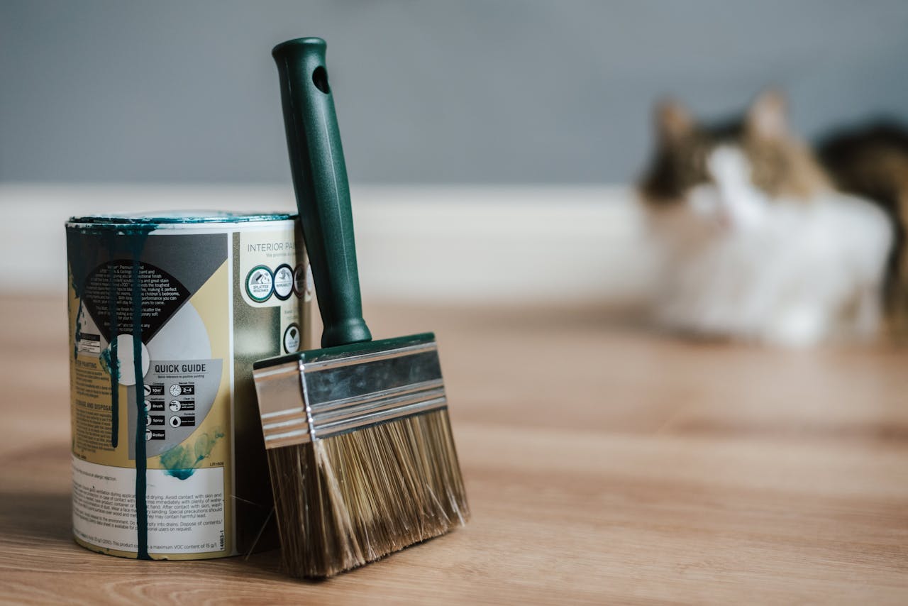 Close-up of paint can and brush on floor with blurred cat in background.